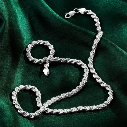 A silver rope-style chain and matching bracelet displayed on a deep green satin fabric. The twisted links create a textured, reflective surface, and both pieces have lobster-claw clasps. Soft highlights and shadows emphasize the draped fabric and the shine of the metal.