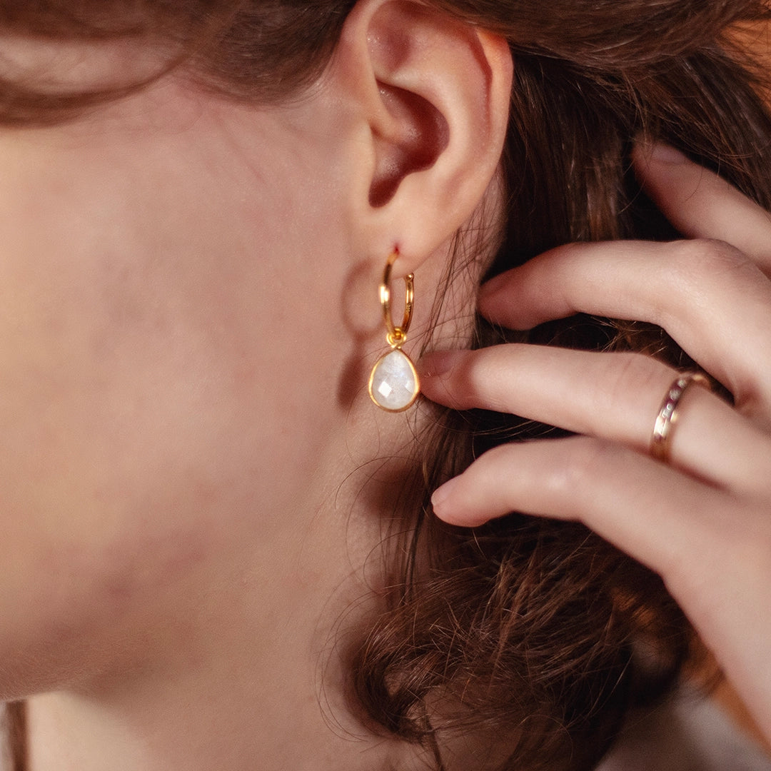 Close-up of a woman wearing a gold hoop earring with a teardrop-shaped moonstone pendant, gently touching it with her hand