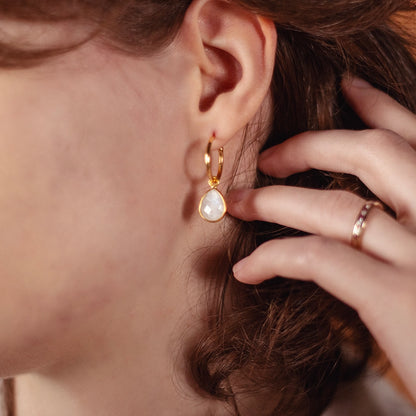 Close-up of a woman wearing a gold hoop earring with a teardrop-shaped moonstone pendant, gently touching it with her hand