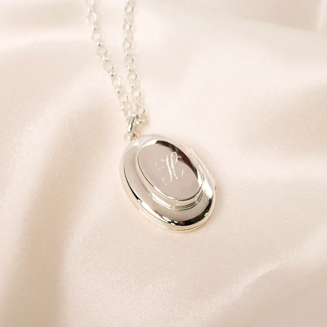 Sterling silver oval locket necklace engraved with the letter “H”, photographed on soft cream fabric with fine silver chain.