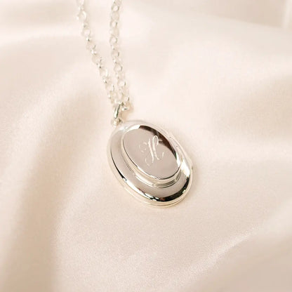 Sterling silver oval locket necklace engraved with the letter “H”, photographed on soft cream fabric with fine silver chain.