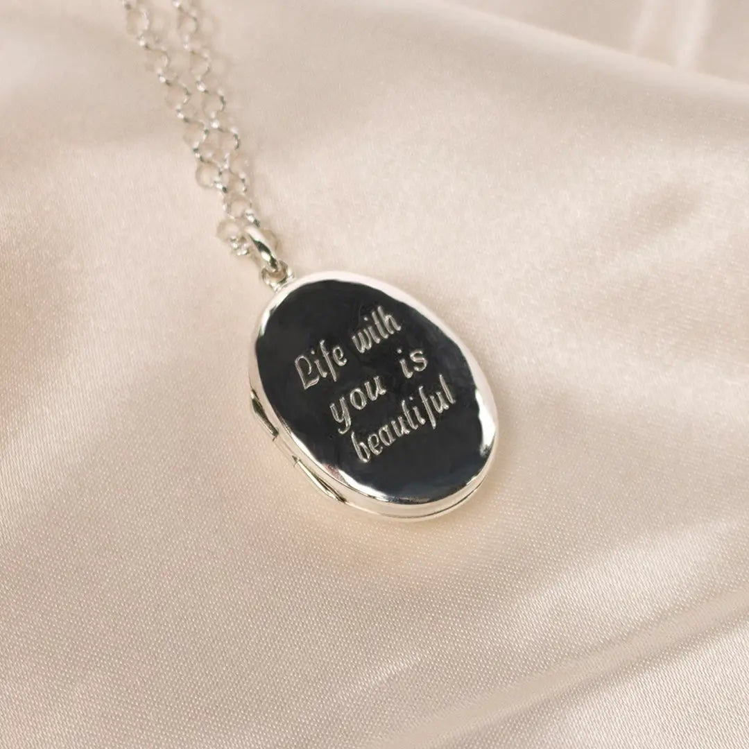 Sterling silver oval locket necklace engraved with the words “Life with you is beautiful”, displayed on soft cream fabric.