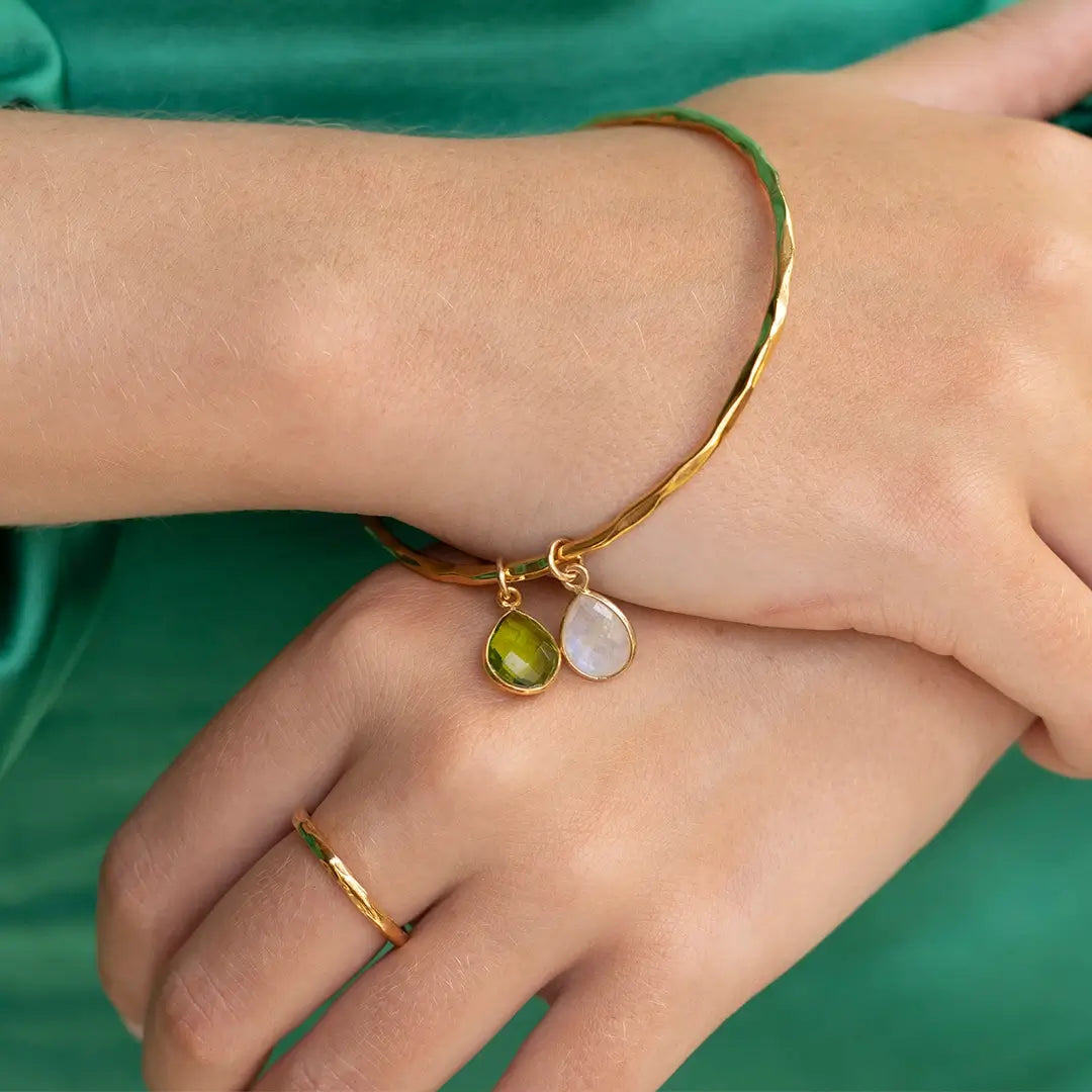 Gold hammered bangle with peridot and moonstone teardrop charms, worn on wrist against a green fabric background, paired with a matching gold ring