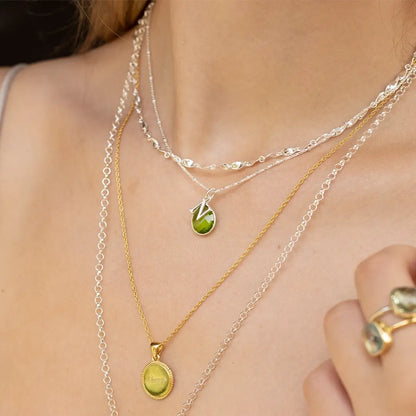 Sterling silver peridot initial necklace featuring a faceted green peridot gemstone, layered with silver chains and a gold coin pendant.