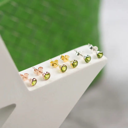 Peridot gemstone stud earrings in sterling silver, gold vermeil and rose gold finishes displayed on a white stand, featuring round green August birthstones.