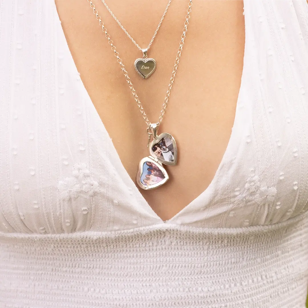 Close-up of layered silver necklaces, including a heart-shaped locket engraved with the word “Love” and two larger heart lockets that open to reveal small personal photos inside. The jewellery is styled with a white textured dress featuring a deep V-neckline.
