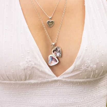 Close-up of layered silver necklaces, including a heart-shaped locket engraved with the word “Love” and two larger heart lockets that open to reveal small personal photos inside. The jewellery is styled with a white textured dress featuring a deep V-neckline.