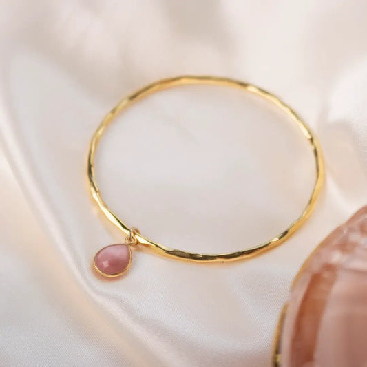 Gold hammered bangle with pink opal teardrop charm resting on soft white fabric, highlighting the gentle blush tone of the gemstone