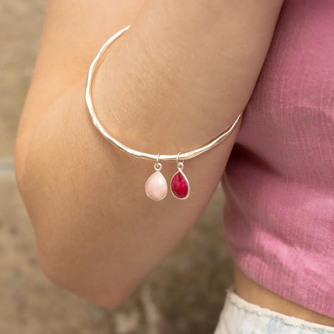 Sterling silver hammered bangle with pink opal and ruby teardrop charms, worn on wrist against a soft neutral and pink fabric background