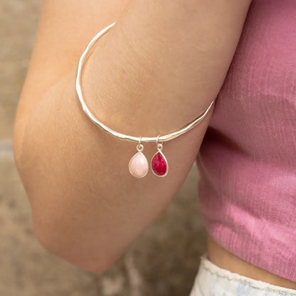 Sterling silver hammered bangle with pink opal and ruby teardrop charms, worn on wrist against a soft neutral and pink fabric background