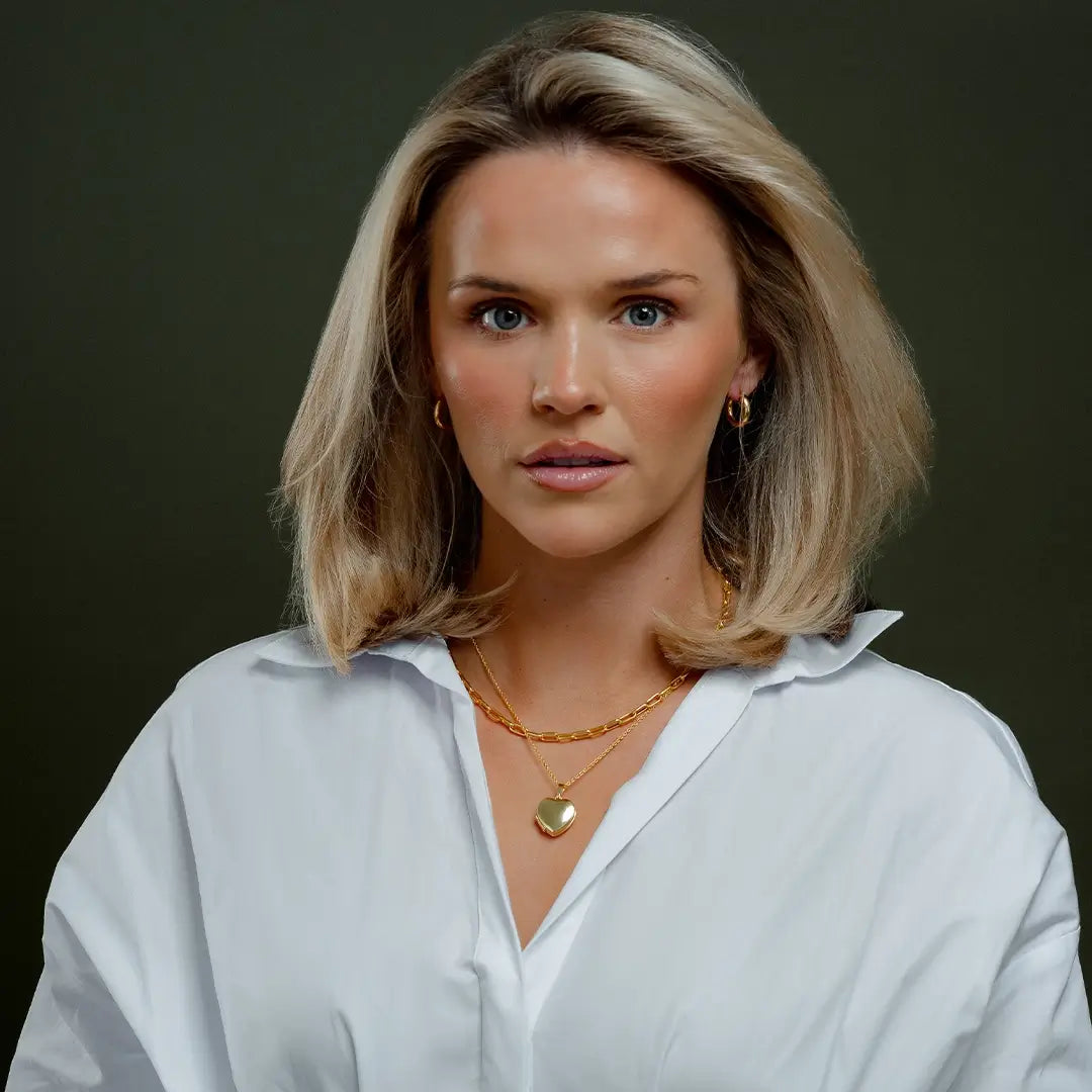 Model wearing a white blouse, gold hoop earrings, and layered gold necklaces featuring a polished gold heart-shaped photo locket. The locket hangs from a fine chain and is shown closed against a dark green background, highlighting its smooth, puffed design and luxurious gold finish.