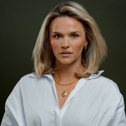 Model wearing a white blouse, gold hoop earrings, and layered gold necklaces featuring a polished gold heart-shaped photo locket. The locket hangs from a fine chain and is shown closed against a dark green background, highlighting its smooth, puffed design and luxurious gold finish.