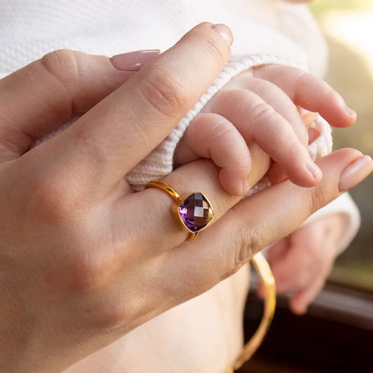 Gold ring with faceted amethyst gemstone worn on hand holding baby’s hand in soft natural light