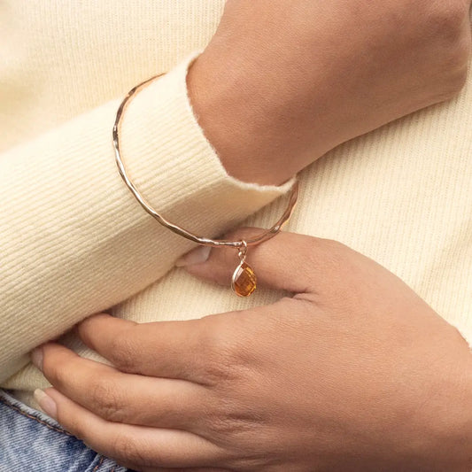 Rose gold hammered bangle with citrine teardrop charm, worn on wrist with a soft cream knit sleeve, hand gently holding the bracelet