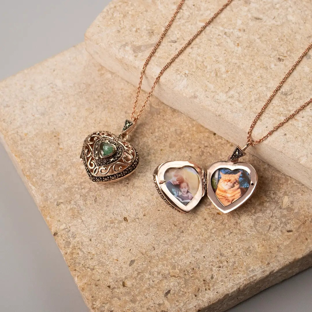 Rose gold vintage heart locket necklaces displayed on stone, one closed with an emerald centre stone and one opened showing personalised photos inside.