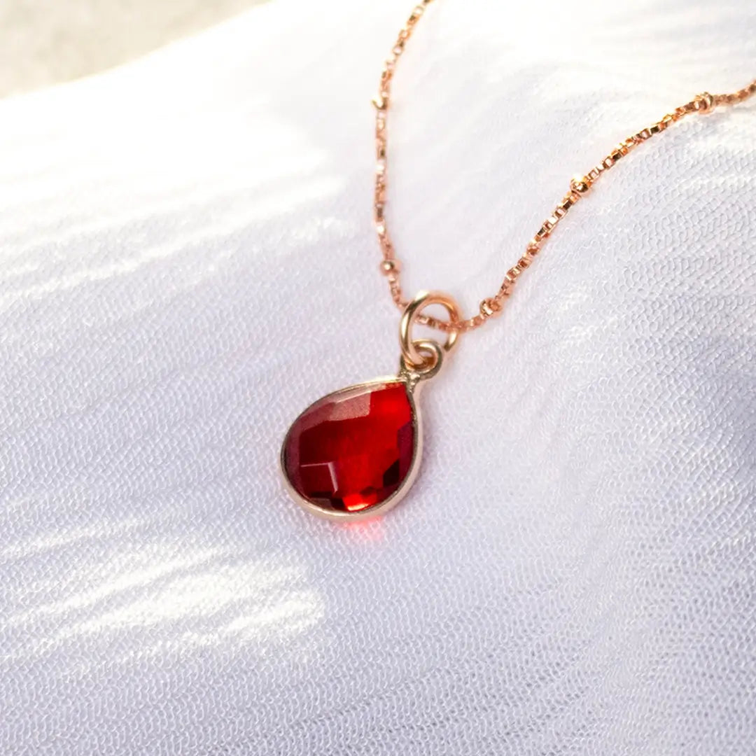 Rose gold vermeil necklace featuring a faceted deep red teardrop gemstone pendant, delicately suspended on a fine chain against a soft white fabric background.