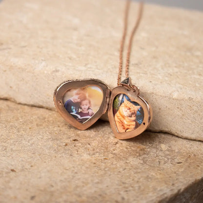 Rose gold heart locket necklace opened to reveal two personalised photo inserts, resting on a stone surface.