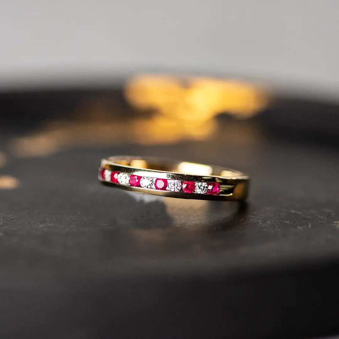 A close-up of a gold band ring set with alternating small round red rubies and clear diamonds, resting on a dark surface with a warm blurred background.