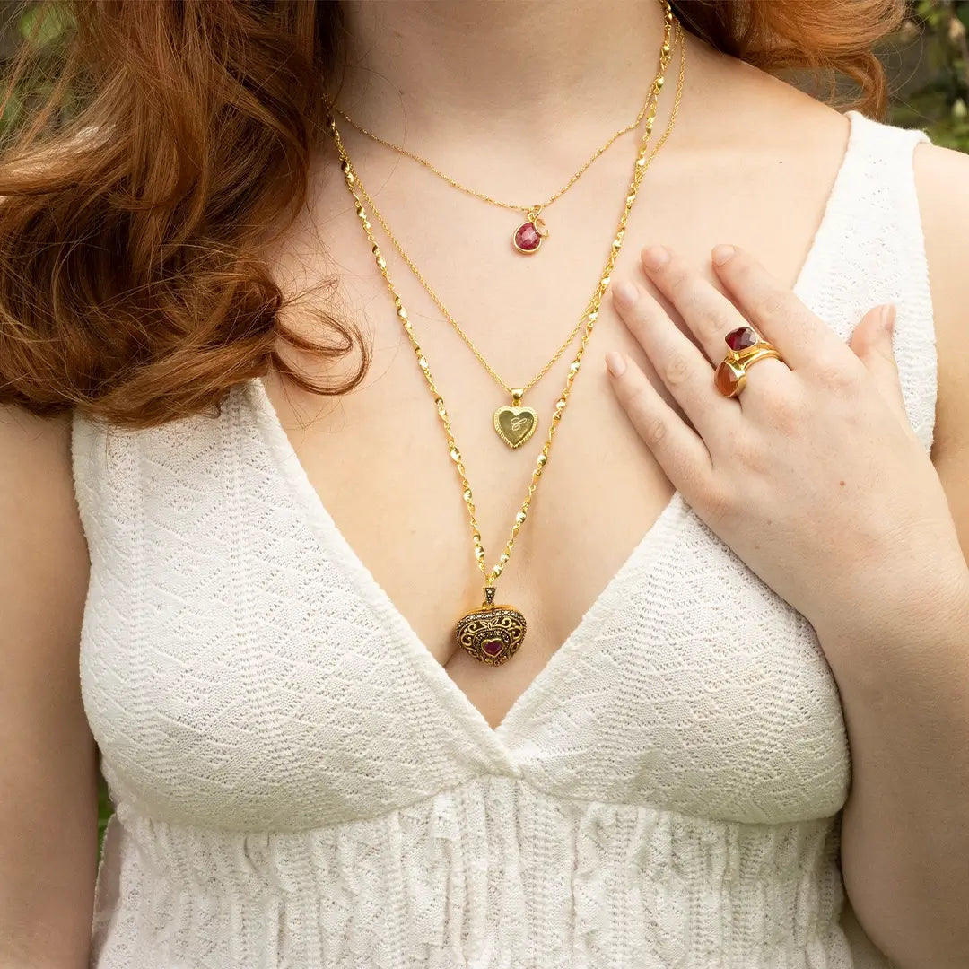 Layered gold necklaces featuring a vintage-style ruby heart locket and ruby pendant, worn with a matching ruby ring on a white dress.