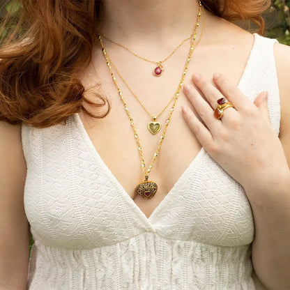 Layered gold necklaces featuring a vintage-style ruby heart locket and ruby pendant, worn with a matching ruby ring on a white dress.