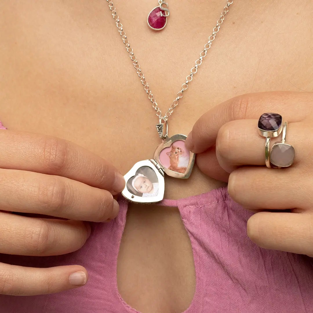 White gold heart locket necklace opened to reveal two personalised photo inserts, shown being held by the wearer, with ruby pendant layered above.