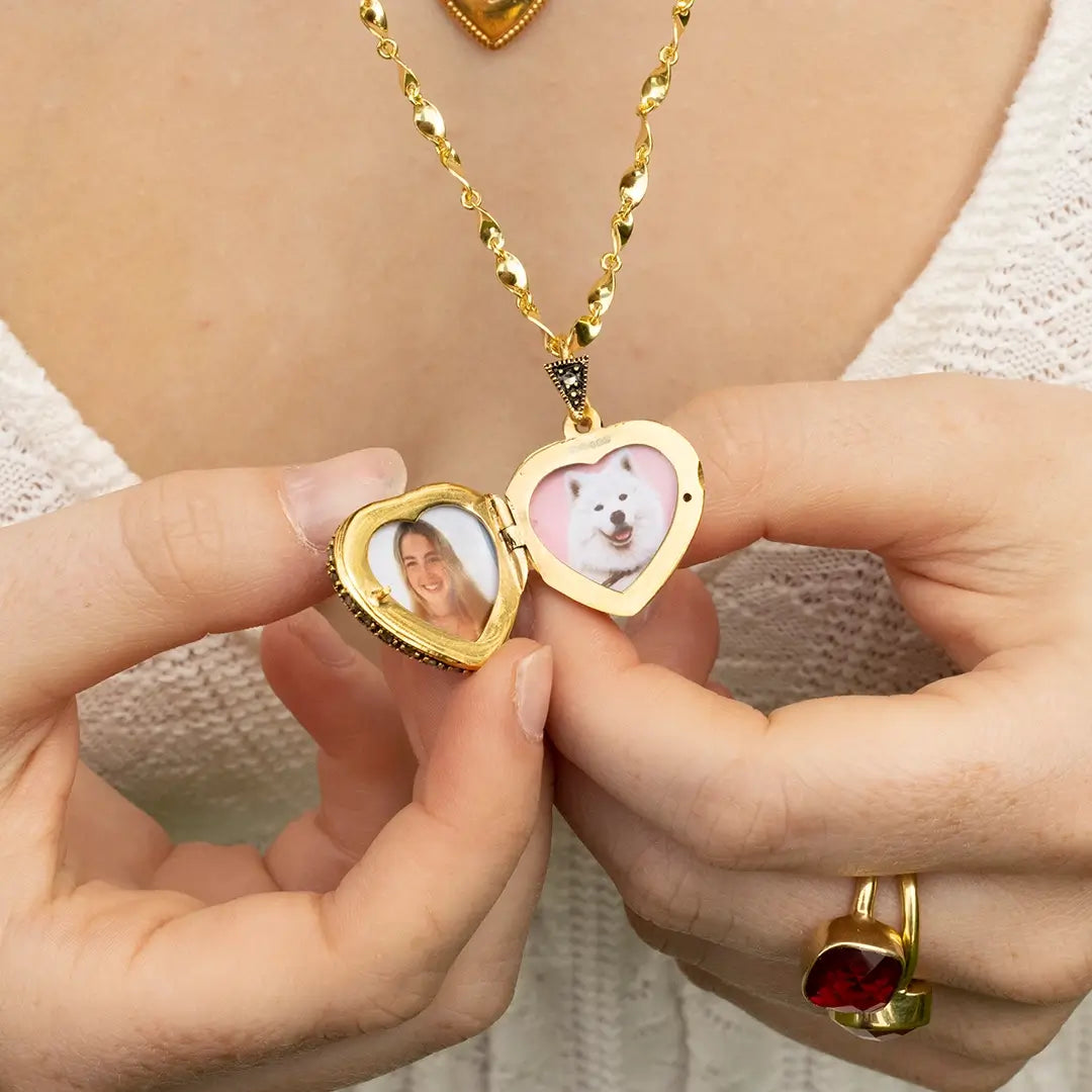 Open gold heart photo locket necklace held by hand, displaying personalised portrait and pet photographs inside, worn on a cream knit top.