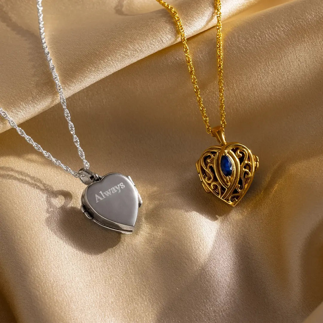 Sterling silver heart locket engraved with “Always” alongside an ornate gold heart locket featuring a blue sapphire centre, both styled on champagne satin.