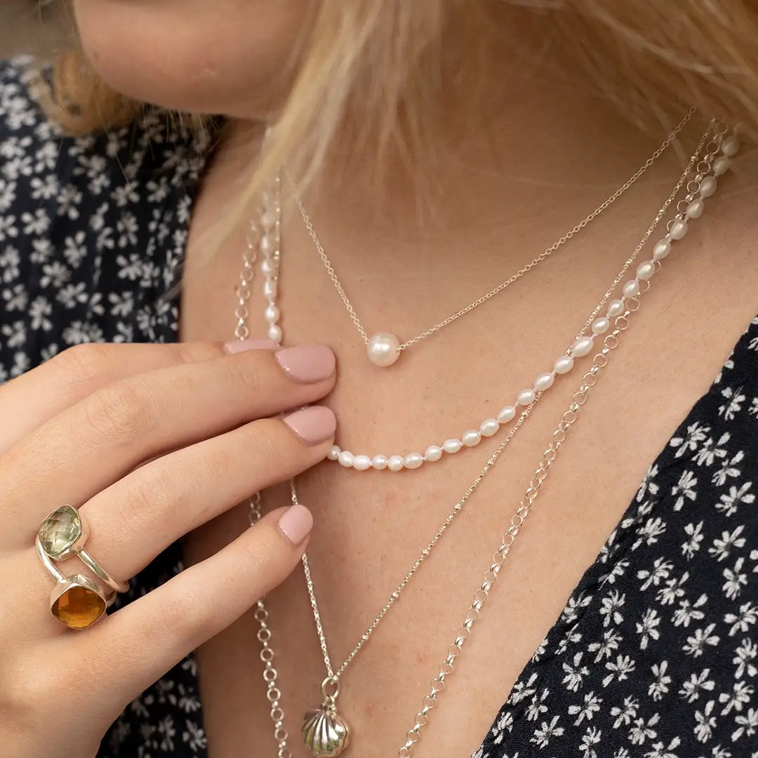 Close-up of layered ivory seed pearl necklace styled with silver chains and pendants, featuring delicate freshwater pearls worn with gemstone rings and a floral print dress.