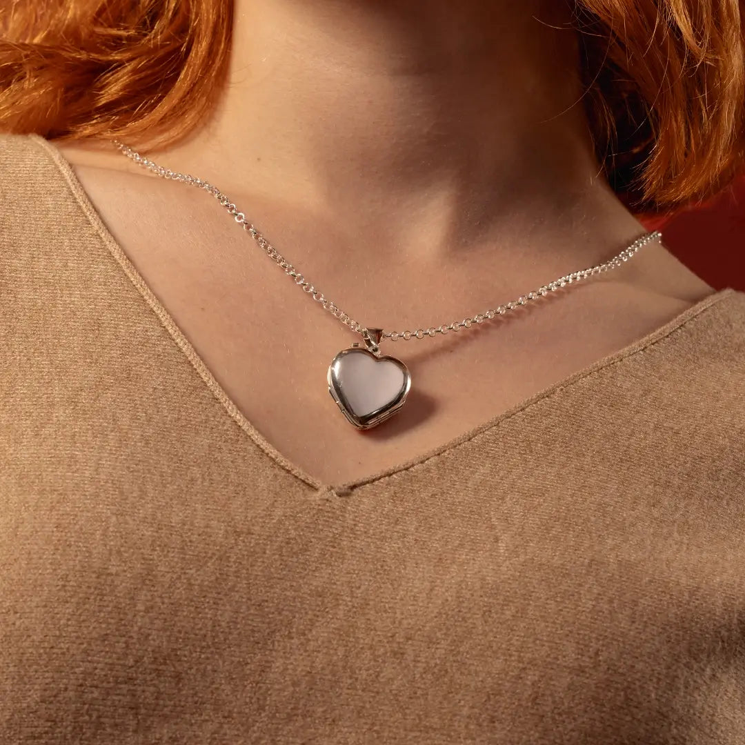 A close-up of a person wearing a delicate silver chain necklace with a polished silver heart-shaped pendant, styled with a brown knit top.