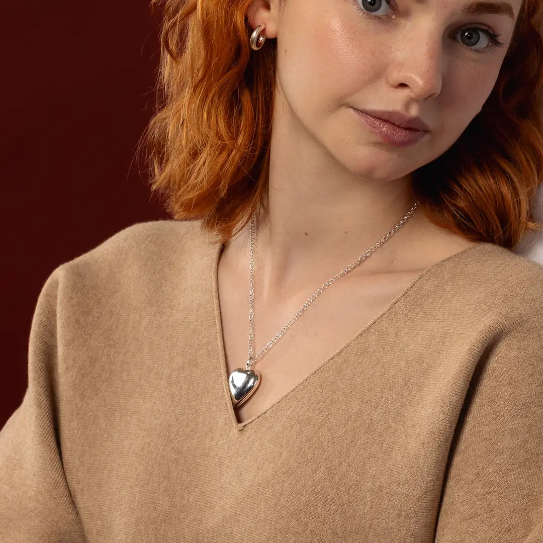 A close-up of a woman with red hair wearing a silver heart-shaped pendant necklace and a small hoop earring, dressed in a beige knit sweater against a dark red background.