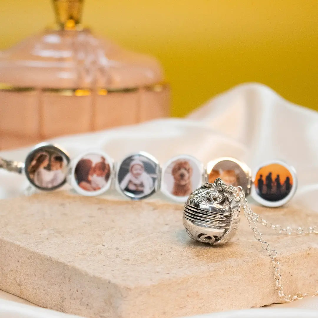 Closed silver Memory Keeper locket and chain on pale stone, with an unfolded six-photo locket in the background.