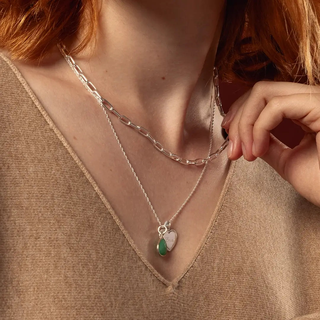 A close-up of a person wearing layered necklaces: a silver paperclip chain necklace and a delicate silver chain with a pendant featuring a silver heart and a chrysoprase stone.