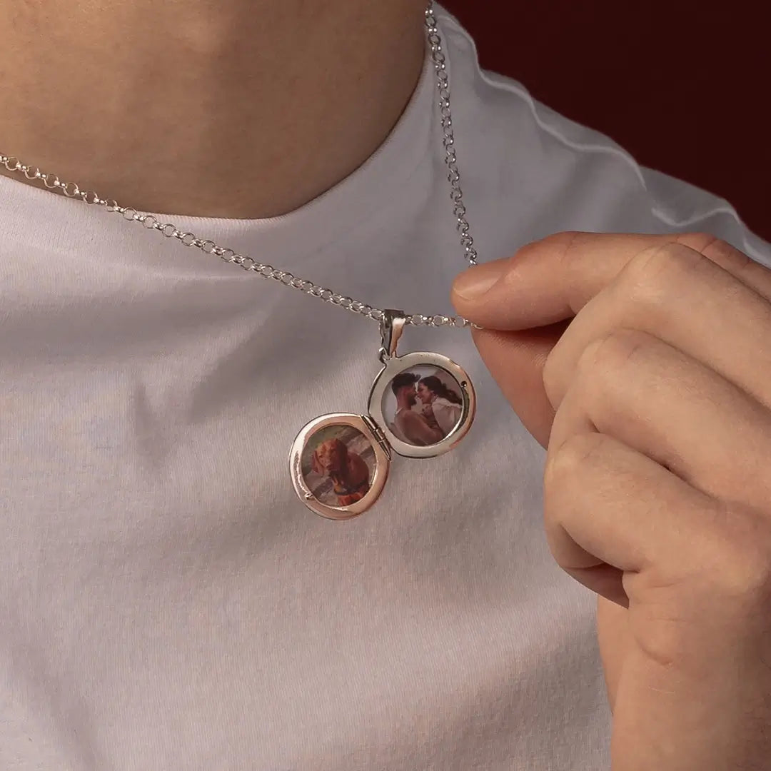 A close-up of a man wearing a silver chain necklace with a round locket pendant that is open to reveal two custom photos inside, while a hand gently holds one side of the locket.