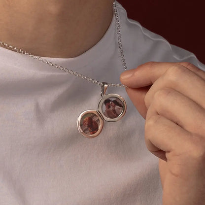 A close-up of a man wearing a silver chain necklace with a round locket pendant that is open to reveal two custom photos inside, while a hand gently holds one side of the locket.