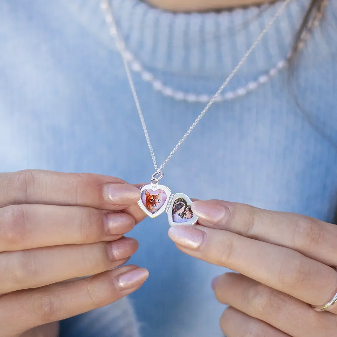 Close-up of a woman’s hands holding an open silver heart-shaped locket necklace, revealing two tiny printed photos inside — one of a cat and one of a couple. She wears the necklace over a light blue sweater with a pearl choker.