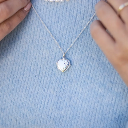 Close-up of a silver heart-shaped locket necklace engraved with a small star detail, styled with a pearl choker over a light blue sweater.
