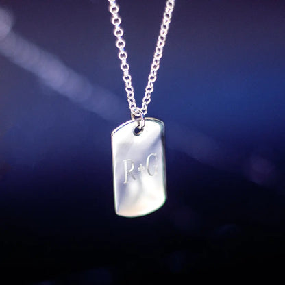 A silver dog tag pendant engraved with the letters “R + G,” hanging from a silver chain against a dark blue gradient background.