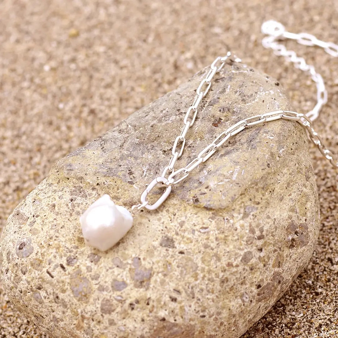 Sterling silver paperclip chain necklace with a single ivory baroque freshwater pearl pendant, displayed draped over a smooth beach stone on sand in natural sunlight.