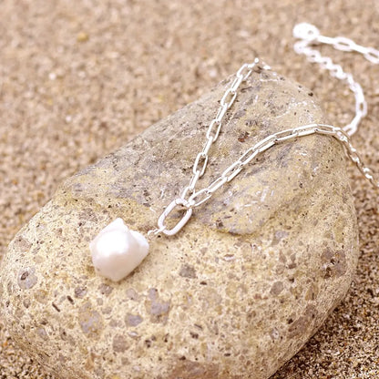 Sterling silver paperclip chain necklace with a single ivory baroque freshwater pearl pendant, displayed draped over a smooth beach stone on sand in natural sunlight.
