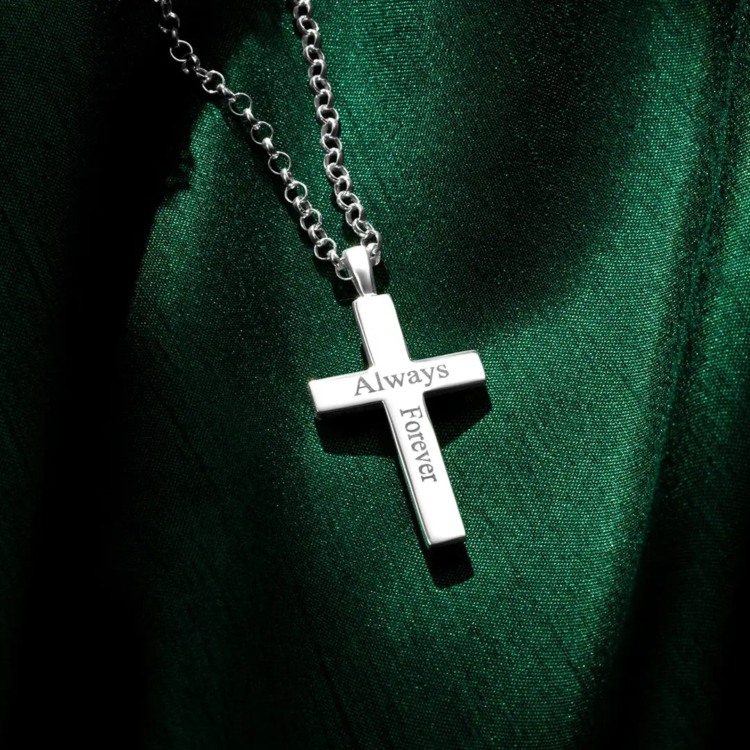 A silver cross pendant engraved with the words “Always” and “Forever,” hanging from a silver chain and resting on rich green fabric with contrasting highlights and shadows.