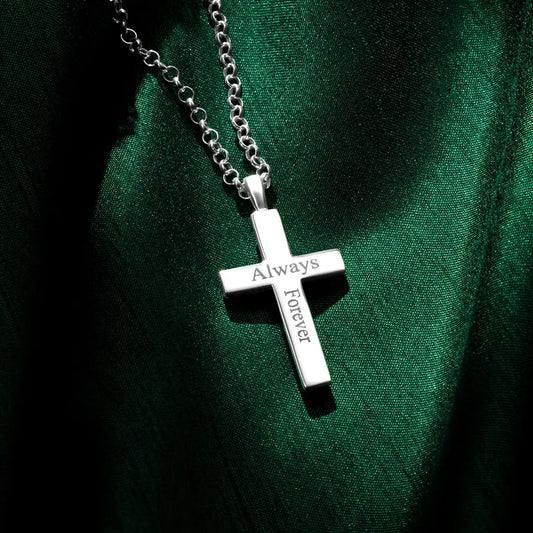 A silver cross pendant engraved with the words “Always” and “Forever,” hanging from a silver chain and resting on rich green fabric with contrasting highlights and shadows.