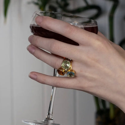 Hand holding a glass of red wine, wearing a gold cocktail ring set with green amethyst and warm-toned gemstones.