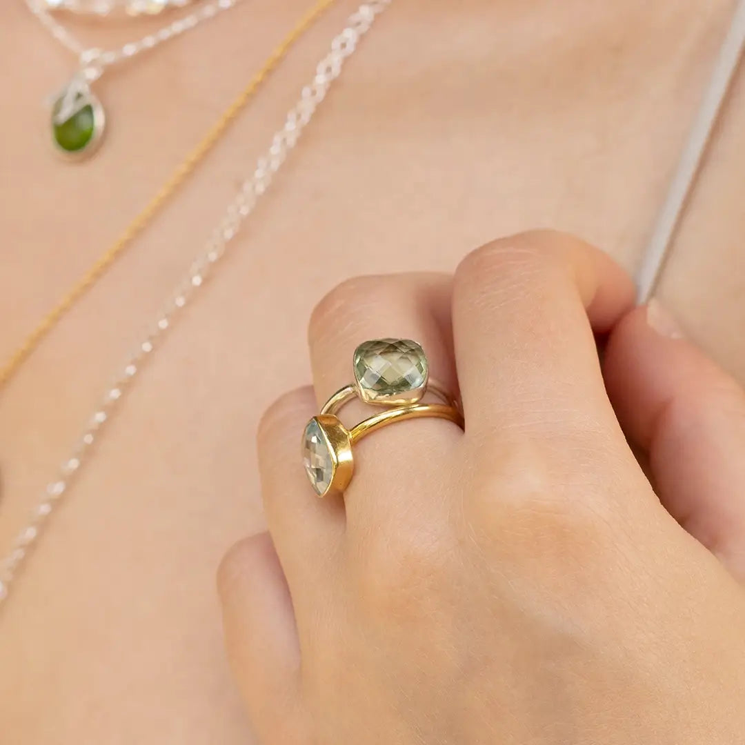 Close-up of a silver green amethyst ring worn on the hand, with layered gold and silver necklaces in the background.