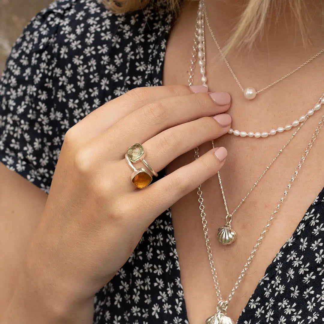 Woman wearing a silver green amethyst ring with a second gemstone, styled with layered pearl and silver necklaces and a patterned dress.