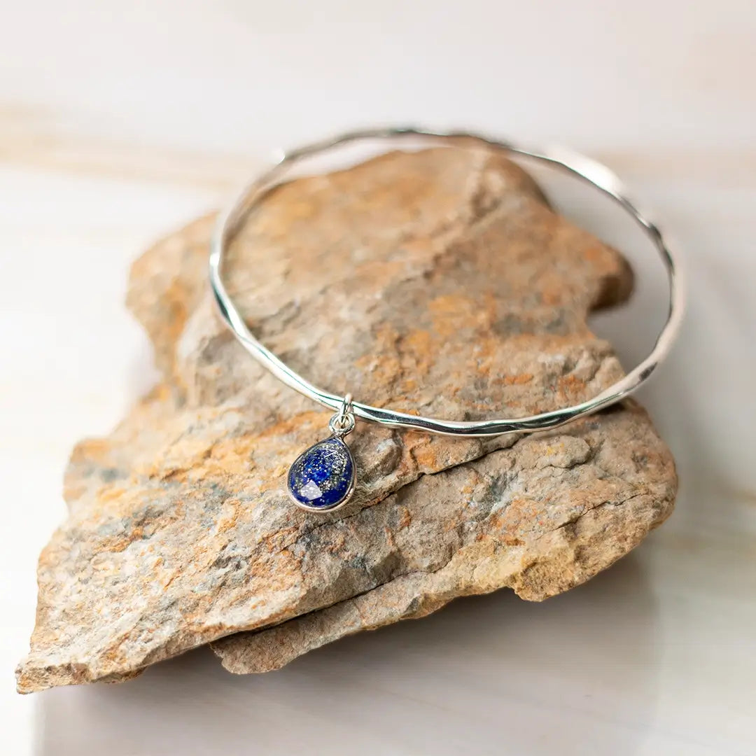 Sterling silver hammered bangle with lapis lazuli teardrop charm, resting on natural stone, highlighting the deep blue gemstone with gold flecks