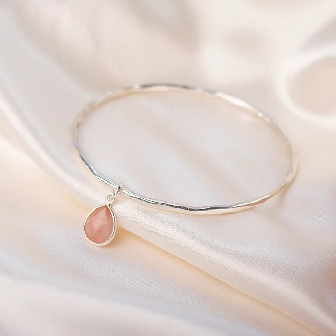 Sterling silver hammered bangle with faceted pink opal teardrop charm resting on soft white satin, highlighting the gentle blush gemstone