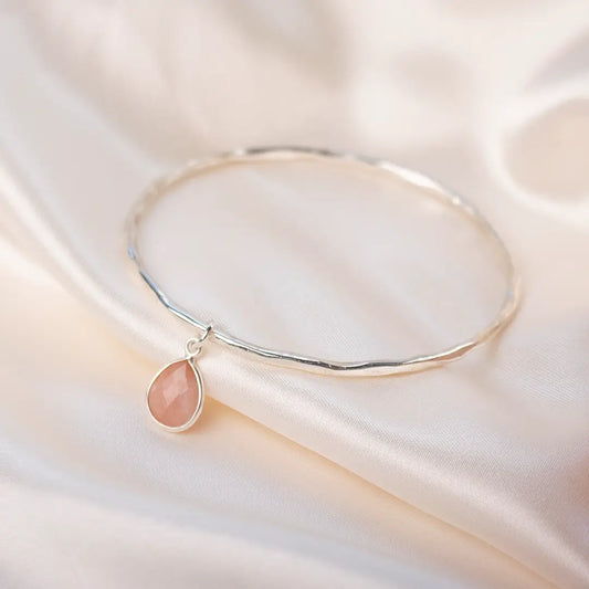 Sterling silver hammered bangle with faceted pink opal teardrop charm resting on soft white satin, highlighting the gentle blush gemstone