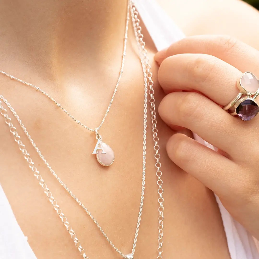 Close-up of layered silver chain necklaces, including a teardrop-shaped pink opal pendant with an initial “A” charm. The woman also wears a large silver ring with purple and pink gemstone settings.