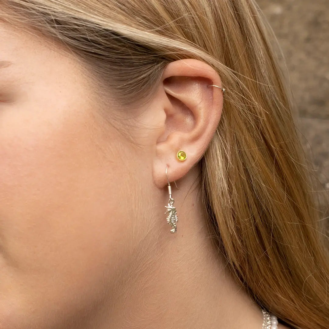 Close-up of a woman’s ear wearing silver seahorse drop earrings and a small round green gemstone stud, with a delicate cartilage piercing.

