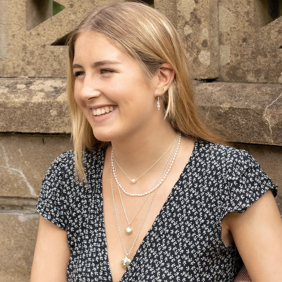 Smiling woman wearing silver seahorse drop earrings and layered silver and pearl necklaces, photographed against a stone wall background.